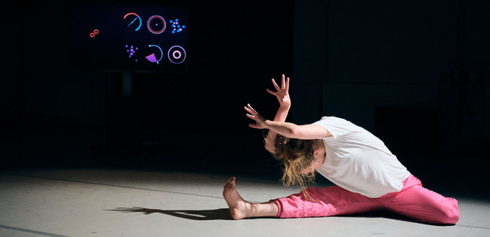 A dancer doing a half split on the floor bows her head and holds up her hands, fingers splayed. Behind, colourful geometric shapes are projects on black cloth.