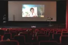 Red theatre seating (mostly empty) rakes down to a wider stage where a zoom call is being projected onto a giant screen. On it, the image of a woman reading from a sheet of paper. 