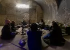 A small circle of people sit in heavy winter coats on the concrete floor of an abandoned building. A nest of microphones sits in the centre of their circle.