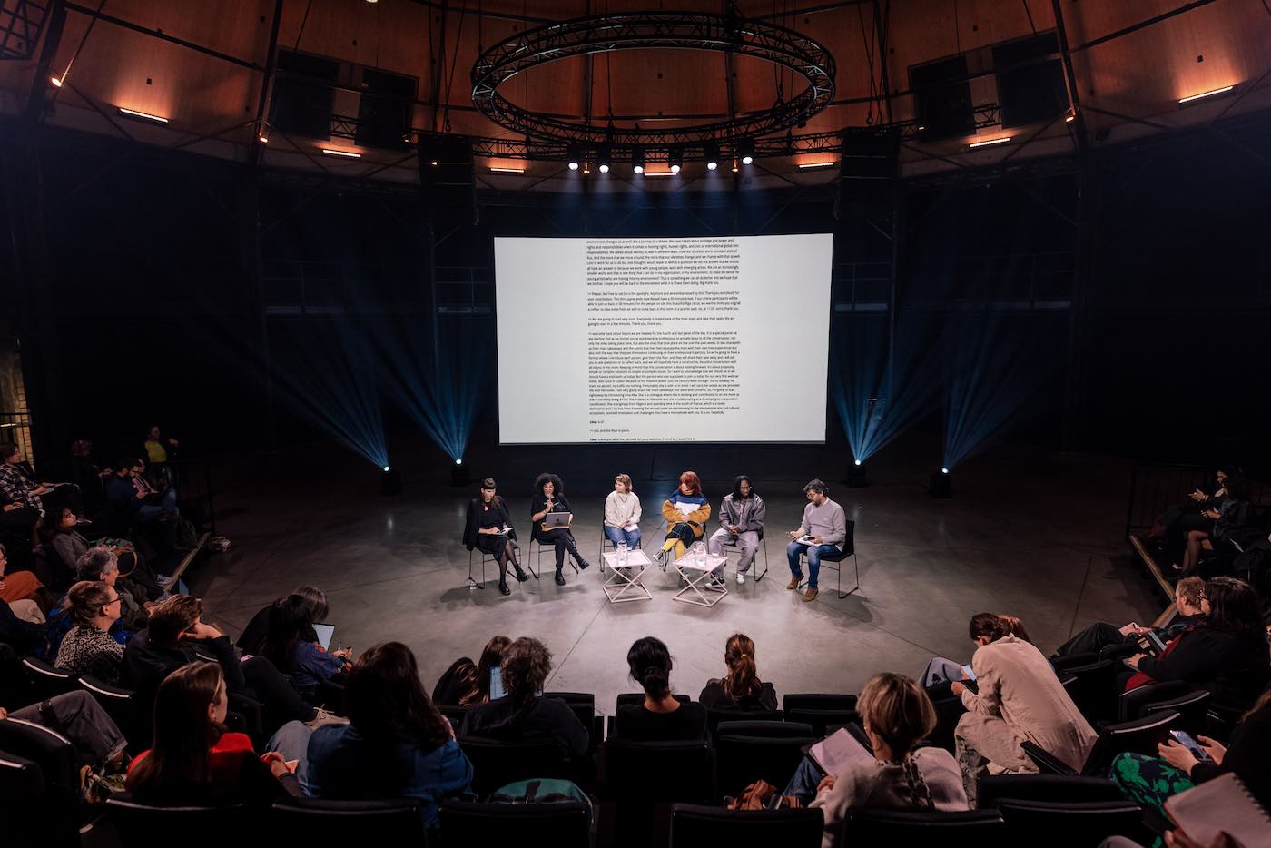 Six people on a stage in front of staggered, semi-circle seating with audience.