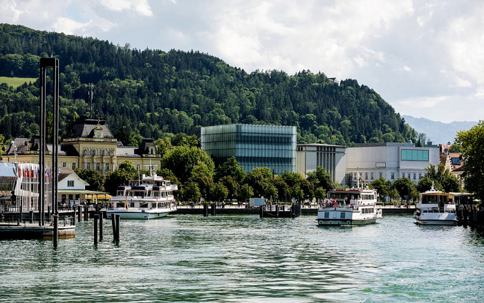 Over the waters of a bay the boxy modernist form of the Kunsthaus Bregenz is visible, nestled in a forested hillside.