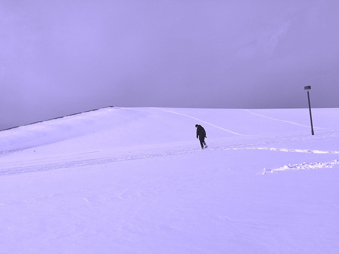 A lone figure trudging across a low hillside covered completely in fresh snow.