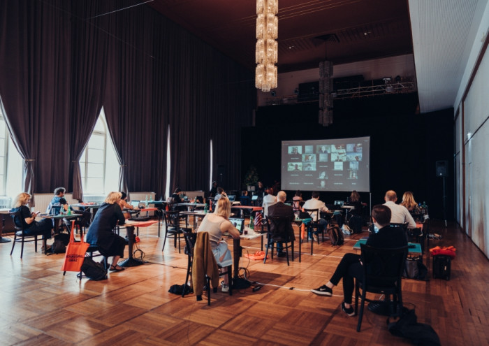 A hybrid digital and live meeting, with the Zoom grid projected on a screen and around a dozen people sitting at individual, socially distanced desks. 