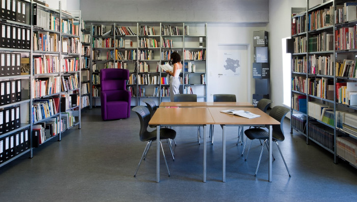 A person in summer clothes reading a book in a room full of bookshelves with two tables and chairs in the centre of the room.