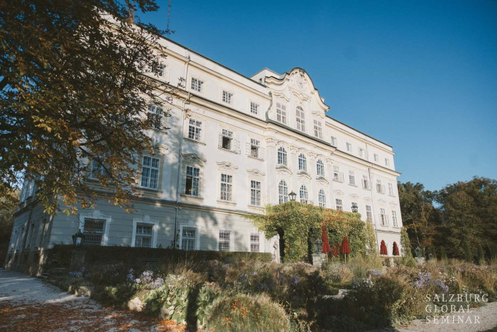 A classical, four-story building surrounded by gardens against a blue sky.
