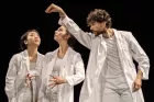 Three dancers all in white. One strikes a pose as if miming a giraffe, one holds an invisible beach ball, the last looks surprised. Choreography is taking place.