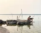 Two artists crouched on a rock at the river's edge. One is scooping something out of the water. Behind them wooden planks and poles are leaned up against each other - makeshift instruments.