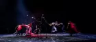 The blurry figures of several circus artists sprinting around three microphones on a stage.