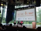 Four speakers on a huge stage in front of a projected powerpoint title screen: 'Music 4 Coop'.