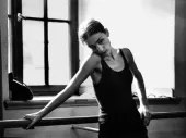 Pina Bausch leans up against a ballet barre, wearing a black vest, giving an appraising look to something out of shot - a dance rehearsal, one can imagine.