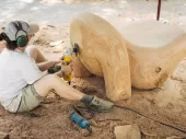 A person sitting on the ground with a grinder carving a wooden sculpture.