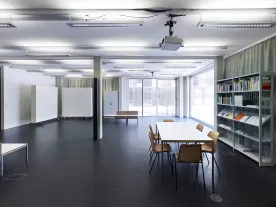 A room with a table and chairs and a bookshelf to the right, windows at the end of the room.
